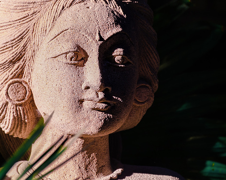 Young woman statue portrait, illuminated by the sun among the palm trees. Bali ancient sculpture, close up. Place for text, copy space.