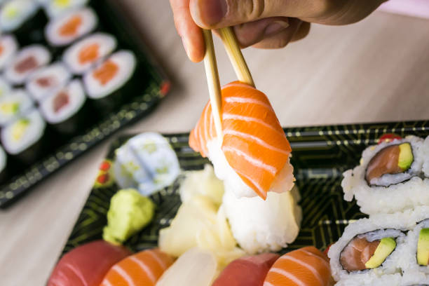 istock Hand using chopsticks pick Sushi and Sashimi rolls on a black stone slatter. Fresh made Sushi set with salmon, prawns, wasabi and ginger. Traditional Japanese cuisine 1082891590