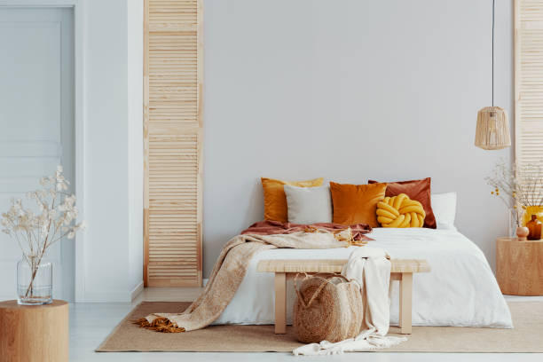 bruine en oranje kussens op witte bed in natuurlijke slaapkamer interieur met rieten lamp en houten nachtkastje met vaas - decor stockfoto's en -beelden