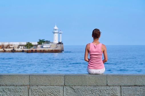 The girl is sitting on the seashore and is looking into the distance to the lighthouse.