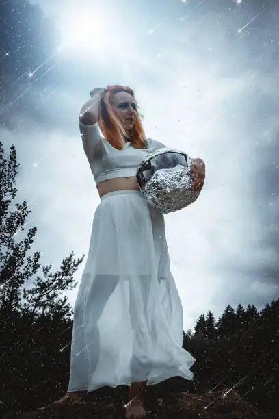 Low Angle Full Length View of Woman Wearing White Dress Standing Holding Helmet on Rocky Cliff Ledge and Looking into the Distance While Stars Falls Around Her in Fantasy Extraterrestrial Themed Image