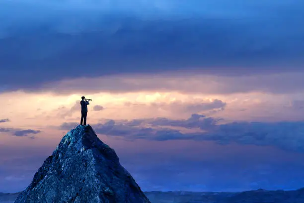 A silhouetted businessman stands on top of a mountain peak and looks into the distance with a spyglass in front of a dramatic sunset in the distance.
