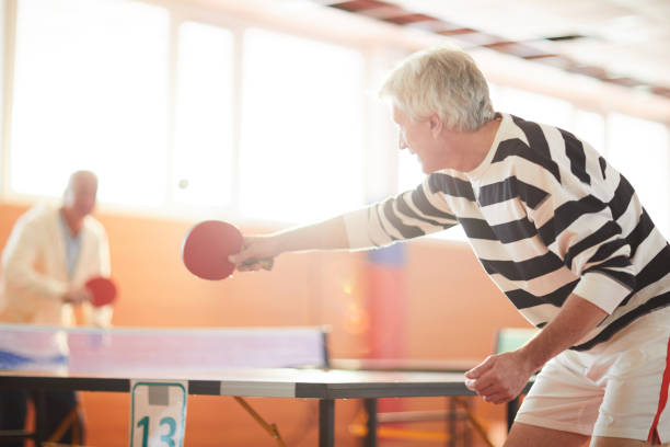 Game of ping pong One of ping pong players hitting ball while passing it over table to his mate during game in the hall tennis senior adult adult mature adult stock pictures, royalty-free photos & images