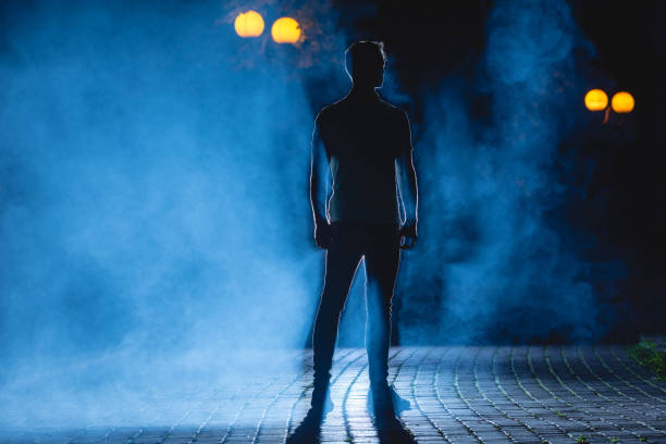 the man stand on the dark alley with a blue fume. night time - street fog profile imagens e fotografias de stock