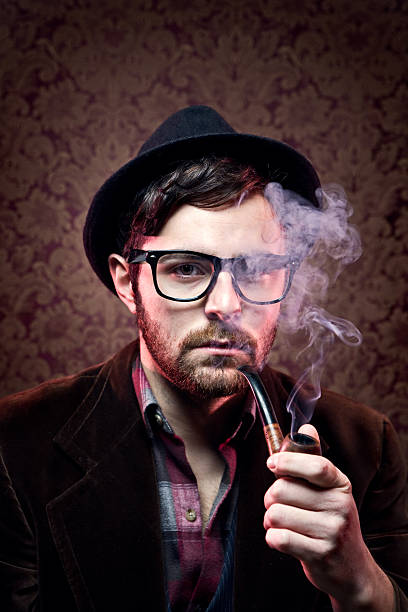 Classy Hipster Portrait A young man with retro frames, fedora hat, and corduroy jacket calmly smokes his pipe while posing for a picture.  Dark Damask patterned wallpaper fills the background behind him.  Vertical with copy space. corduroy jacket stock pictures, royalty-free photos & images