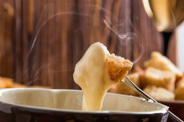 Close up, macro. Traditional french cheese fondue. Crouton dipped into stemy-hot cheese fondue on a long-stemmed fork. Winter seasonal meal. Copy space.