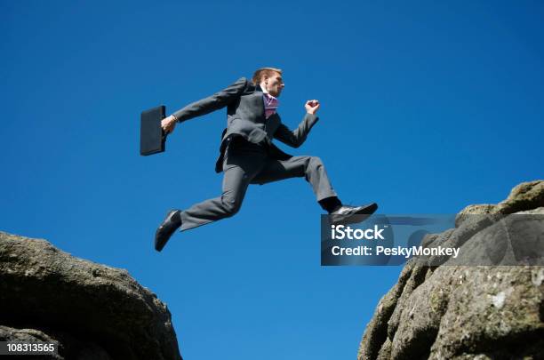 Photo libre de droit de Courageux Homme Daffaires Sauter Entre Rocks Contre Ciel Bleu banque d'images et plus d'images libres de droit de Affaires