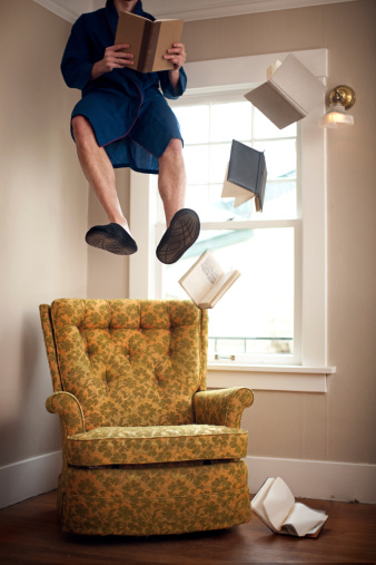 A man floats suspended in mid air as he reads a book in his living room, surrounded by other floating / falling books.  Vertical with copy space.
