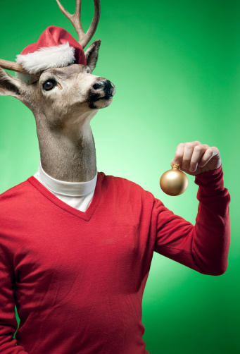 A stag / buck / reindeer dressed in traditional Christmas holiday colors and a Santa Claus hat holds up a bauble ornament in front of a brightly lit green background.  Vertical with copy space.