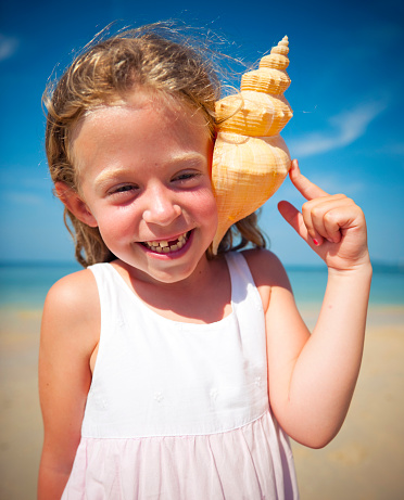 happy trendy woman in white swimsuit on the beach laying on a striped towel and listening to seashell. perfect shell - nice inexpensive souvenir. Sun protected hair. total relaxation on the best beach