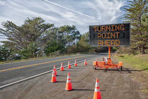 TURNING POINT AHEAD:  This is a photograph of a mobile roadside sign parked on highway one in northern California. It is a trailer and powered by batteries and provides information and warnings for drivers by displaying words on a large panel display.