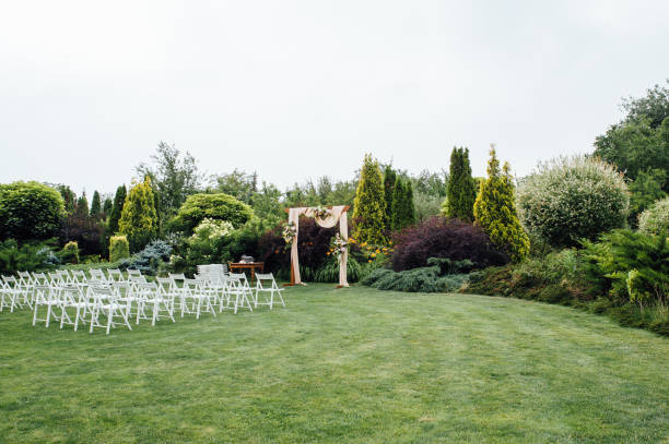 White wedding chairs decorated with fresh flowers on a green grass. White wedding chairs decorated with fresh flowers on a green grass. Empty wooden chairs for guests on green lawn in the garden prepared for wedding ceremony. country wedding arches stock pictures, royalty-free photos & images