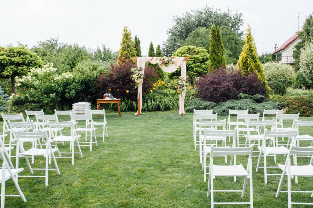 White wedding chairs decorated with fresh flowers on a green grass. White wedding chairs decorated with fresh flowers on a green grass. Empty wooden chairs for guests on green lawn in the garden prepared for wedding ceremony. country wedding arches stock pictures, royalty-free photos & images