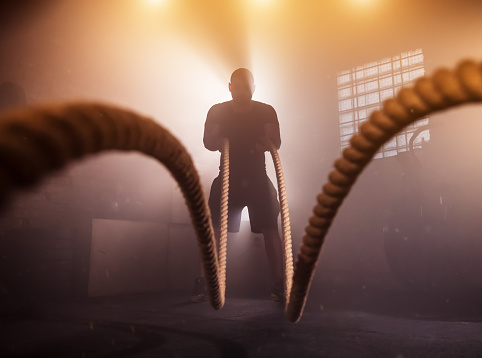 Young man doing hard exercise workout in gym interior with rope waving. Cinematic mood with dust, dramatic lightning and smoke. Active and healthy lifestyle.