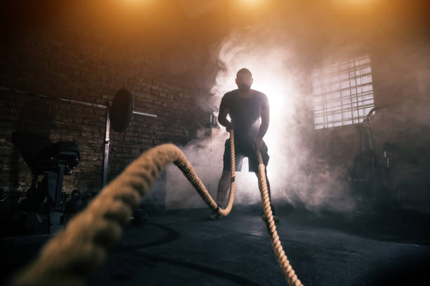 Young man doing hard exercise workout in gym Young man doing hard exercise workout in gym interior with rope waveing. Cinematic mood with dust, dramatic lightning and smoke. Active and healthy lifestyle. toughness stock pictures, royalty-free photos & images