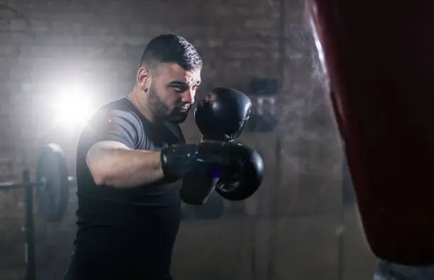 Young man hitting punching bag during training lesson. Dark industrial fitness workout interior with cinematic atmosphere. Active sport and healthy lifestyle.