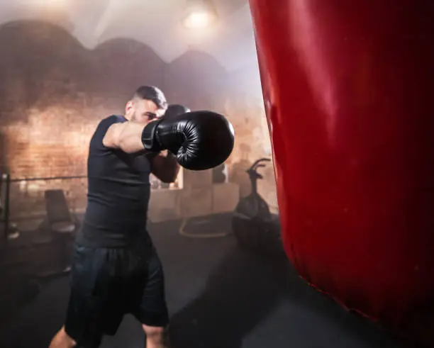 Young man hitting punching bag during training lesson. Dark industrial fitness workout interior with cinematic atmosphere. Active sport and healthy lifestyle.