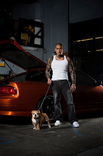 Tough looking guy with tattoos standing in front of his car with a puppy bulldog in a garage. Copy space.