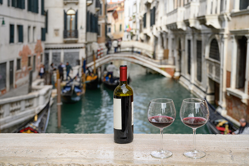 Bottle of red wine with two glasses on marble bridge over canal in Venice, Italy. Shallow DOF with selective focus on the foreground.