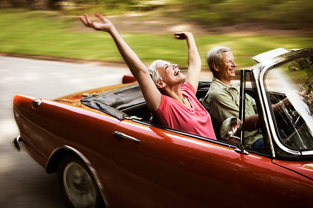 Happy Senior Couple Going For a Drive Senior woman throws her arms in the air while her husband drives her down a road in their convertible. Horizontal shot. collectors car stock pictures, royalty-free photos & images