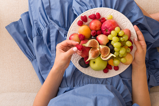 Girl holding white plate with apricots, raspberries, strawberries, figs and grapes on her knees. Top view on woman sitting cozy on the couch at home and enjoying delicious summer fruit and berries.