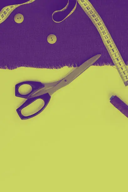 Ultra violet falric, measuring tare, two buttons and cutting-out scissors with black handle as diagonal on bright yellow  background. Top view. copy space.