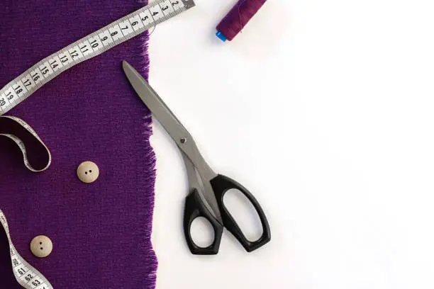 Ultra violet falric, measuring pare, two bullons and cutting-out scissors with black handle as diagonal on the white horizontal background. Top view. copy space.