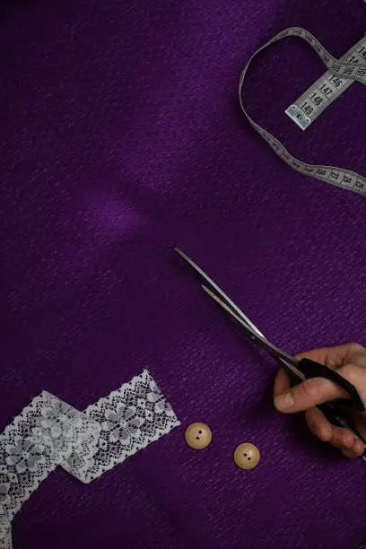 vertical piece of ultraviolet cloth, with measuring tape, buttons, human hands with cutting-out scissors, lace. Cloth is cut diagonally Top view, Copy space,