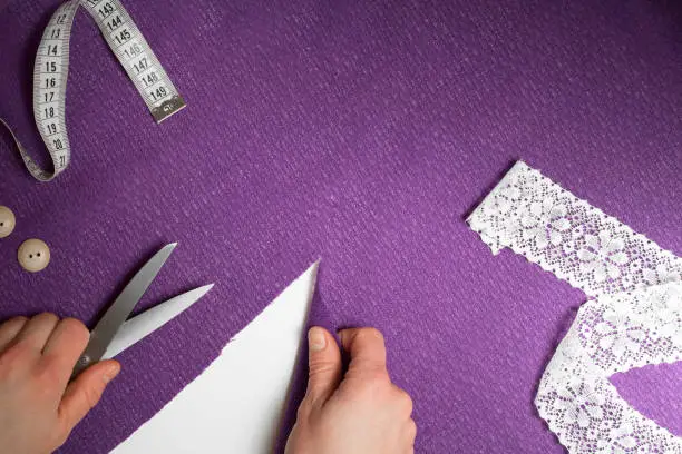 Horizontal piece of ultraviolet cloth, with measuring tape, buttons, human hands with cutting-out scissors. Cloth is cut diagonally, white background under. Top view, Copy space.