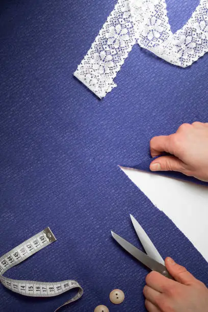 Horizontal piece of blue cloth, with measuring tape, buttons, human hands with cutting-out scissors. Cloth is cut diagonally, white background under. Top view, Copy space.