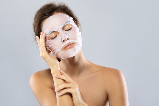 Face care and beauty treatments. Woman with a sheet moisturizing mask on her face isolated on gray background