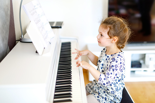 Beautiful little kid girl playing piano in living room or music school. Preschool child having fun with learning to play music instrument. Education, skills concept.