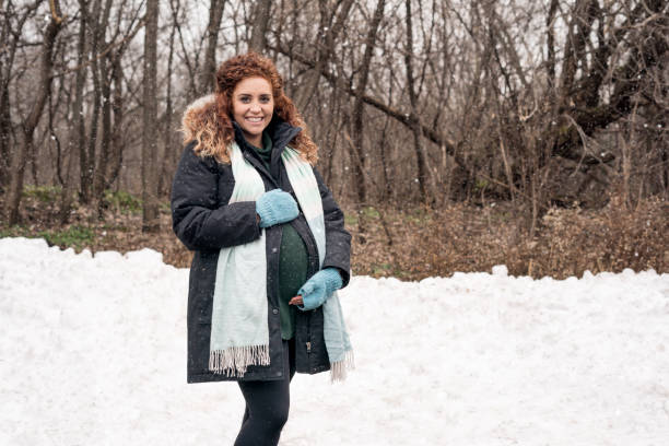 Beautiful Hispanic prenant woman with long curly red auburn hair wearing a green sweater, coat, scard and fingerless gloves standing in snow in a forest in late Autumn, Minnesota