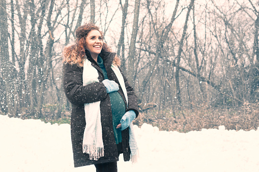 Beautiful Hispanic prenant woman with long curly red auburn hair wearing a green sweater, coat, scard and fingerless gloves standing in snow in a forest in late Autumn, Minnesota