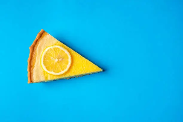 Slice of yellow lemon cake decorated with a slice of lemon, on a blue background. Directly above view. Minimalist style. Delicious fruit dessert.