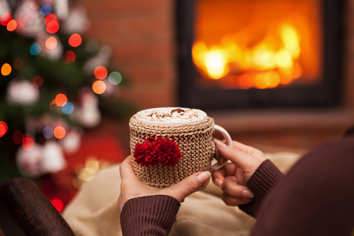 Woman relaxing with a cup of hot chocolate sitting in an armchair by the fireplace and christmas tree - closeup on winter evening relaxation detail