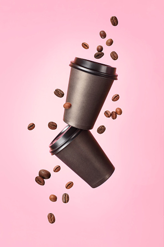 Flying coffee from a paper cups with flying coffee beans on a pink background. Coffee concept. Mock up. Empty polystyrene coffee drinking mug mockup front view. Clear plain tea take away package.