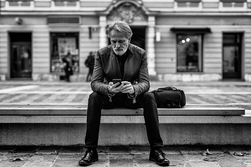 Mature man sitting on a bench in the city and using phone