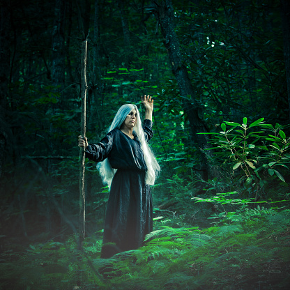woman dress with  a black costume, grey wig and a wooden stick, pretending to make conjures and spell in the woods.