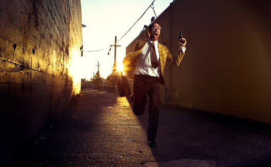 A spy in vintage 1970's clothes runs down a dark city alley yelling with a gun and a briefcase, apparently being chased.  The sun sets brightly behind him.  Horizontal with copy space.  Slight motion blur.