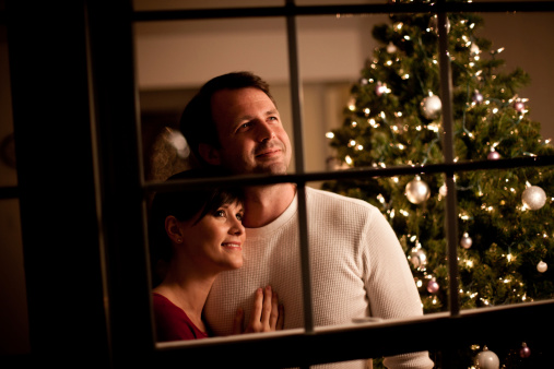 Color photo of a happy couple hugging while looking out the window together at home with Christmas tree and lights behind them. Shot from outside at night. (NOTES: Focus is on the man's face. Grain from higher ISO due to nightime shot and ambient light usage.)