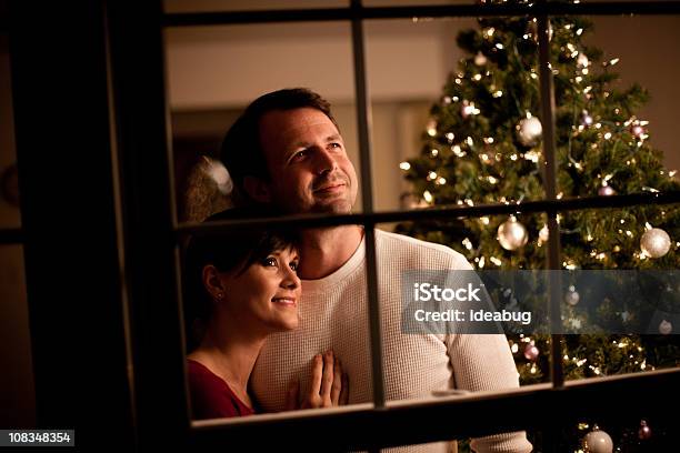 Foto de Casal Feliz Olhando Pela Janela Com Árvore De Natal Atrás e mais fotos de stock de Janela