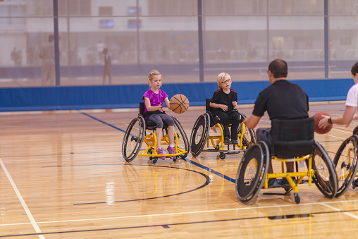 Two kids and two adults are in a gymnasium. They are all using wheelchairs and playing with two basketballs.