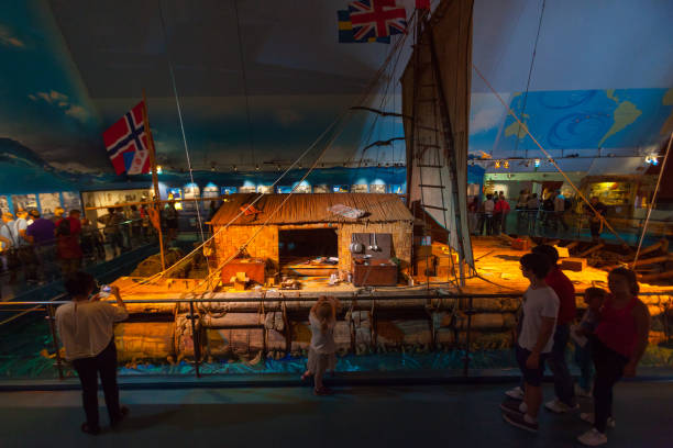 The visitors of the Kon-Tiki Museum are looking at and taking photos of one of the exhibits - a historical household on a float. The Kon-Tiki Museum is a museum in the Bygdoy peninsula in Oslo, Norway. It houses vessels and maps from the Kon-Tiki expedition, as well as a library with about 8000 books. It was opened in 1949.