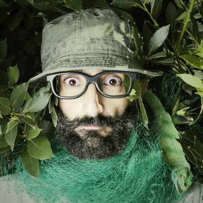 One nerdish looking middle age man looking scared. He is inside some bushes with a lizard on his shoulder. Wearing a green camp hat and black rimmed glasses.