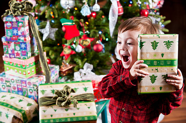 Boy holding gift to ear in front of Christmas tree and gifts stock photo