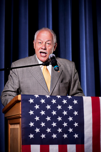 American mature politician in suit standing at tribune and giving a speech during press conference