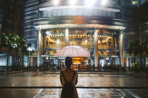 Young woman - solo traveler - enjoying her time in Malaysia, traveling and sightseeing. As the night falls, she is passing by the famous Twin towers, holding the umbrella on a rainy night.