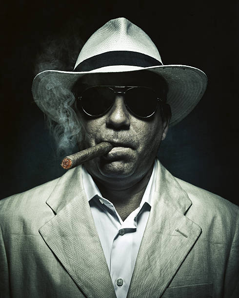elegant cuban mysterious man smoking a cigar tropical elegance (spy, detective or mafia guy) smoking a cigar- mature man wearing hat, summer suit and dark sunglasses on black background. square format mafia boss stock pictures, royalty-free photos & images