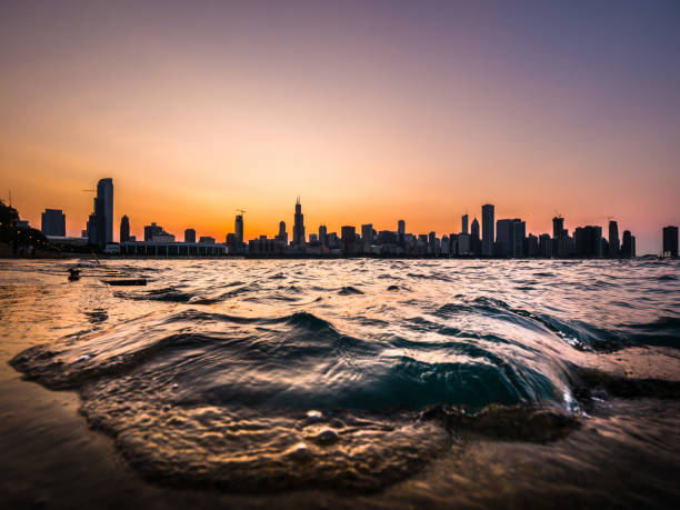 Chicago skyline picture during a beautiful sunset with purple and orange sky above and building silhouettes on the horizon with a wave from Lake Michigan coming up over the concrete shoreline edge. stock photo